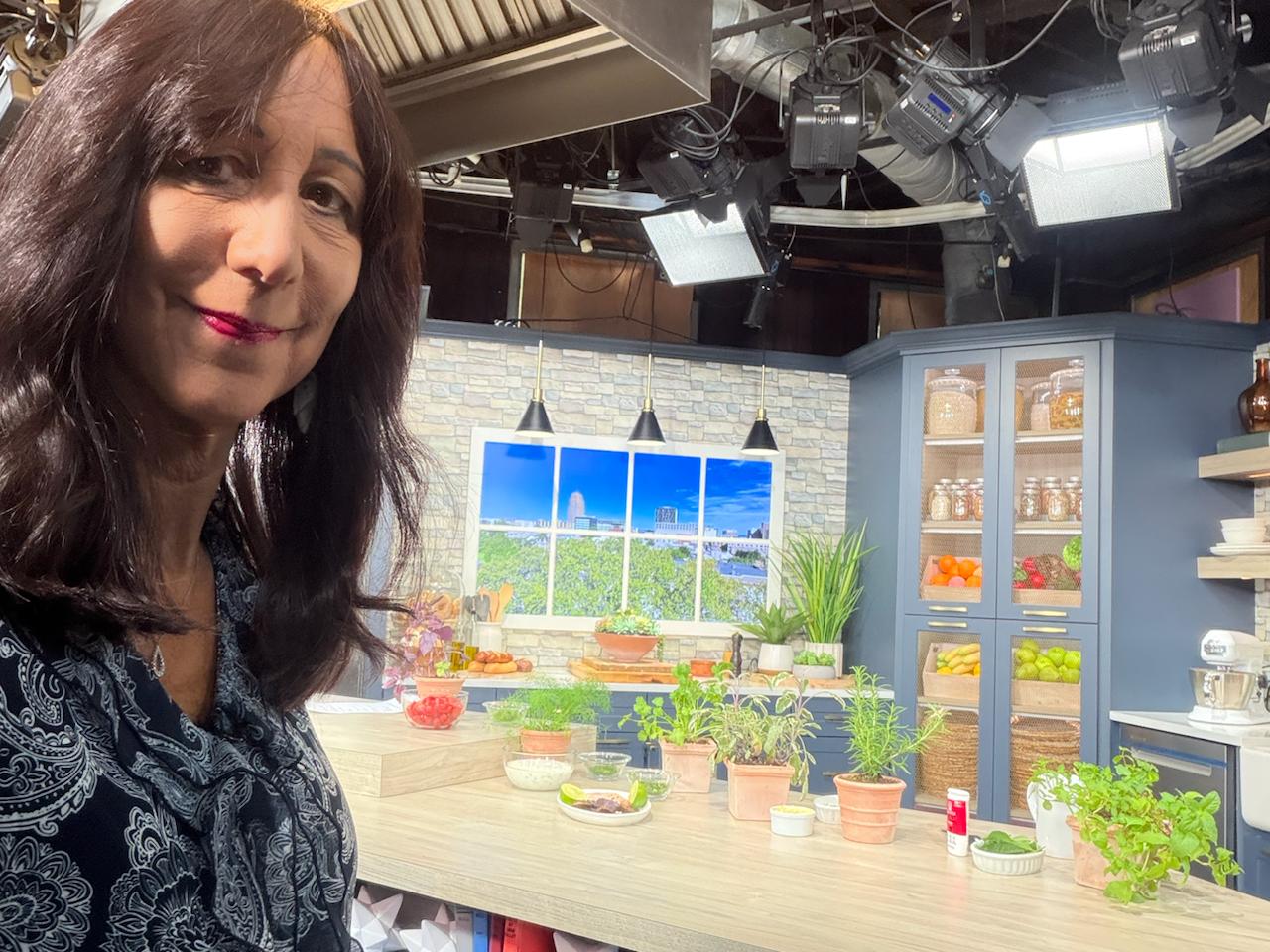 Luisa Szakacs on a WFMZ studio kitchen set with fresh herbs, produce, and cooking ingredients.